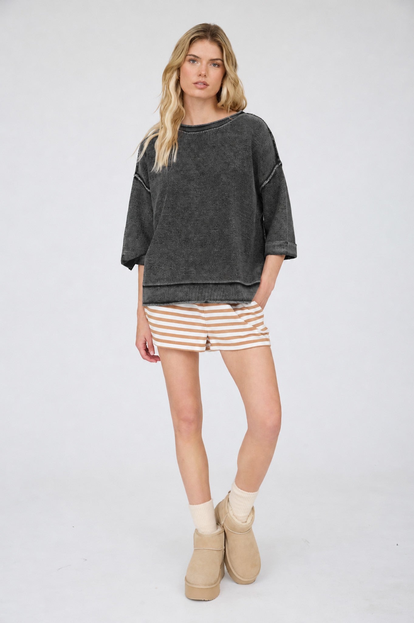 Woman wearing a dark gray oversized sweatshirt, striped shorts, and beige boots on a white background