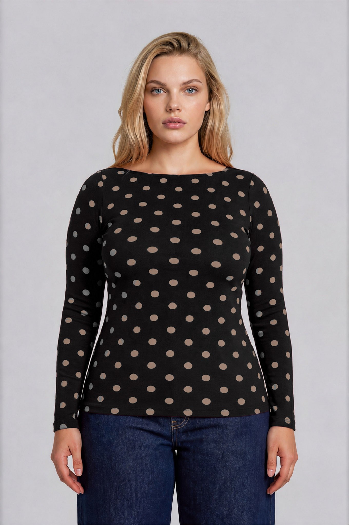 A woman modeling a black long-sleeve knit top with a beige polka dot pattern, paired with dark blue jeans, against a solid light gray background.