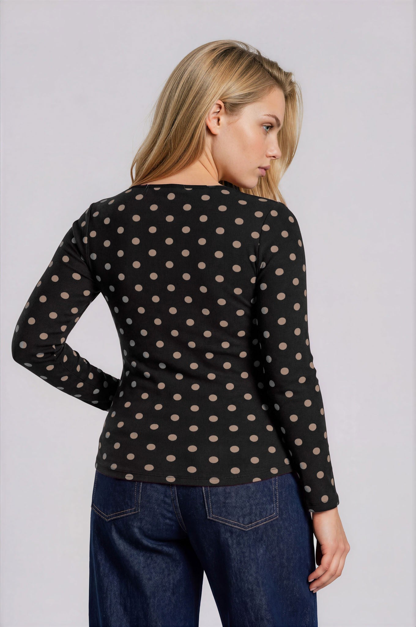 Woman wearing a black polka dot long-sleeve top and blue jeans against a white background