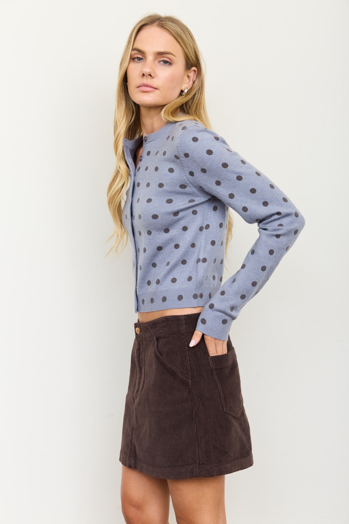 A blonde model wearing a light blue-gray long-sleeve knit top with a dark gray polka dot pattern, paired with a dark brown corduroy mini skirt, against a plain off-white studio background.