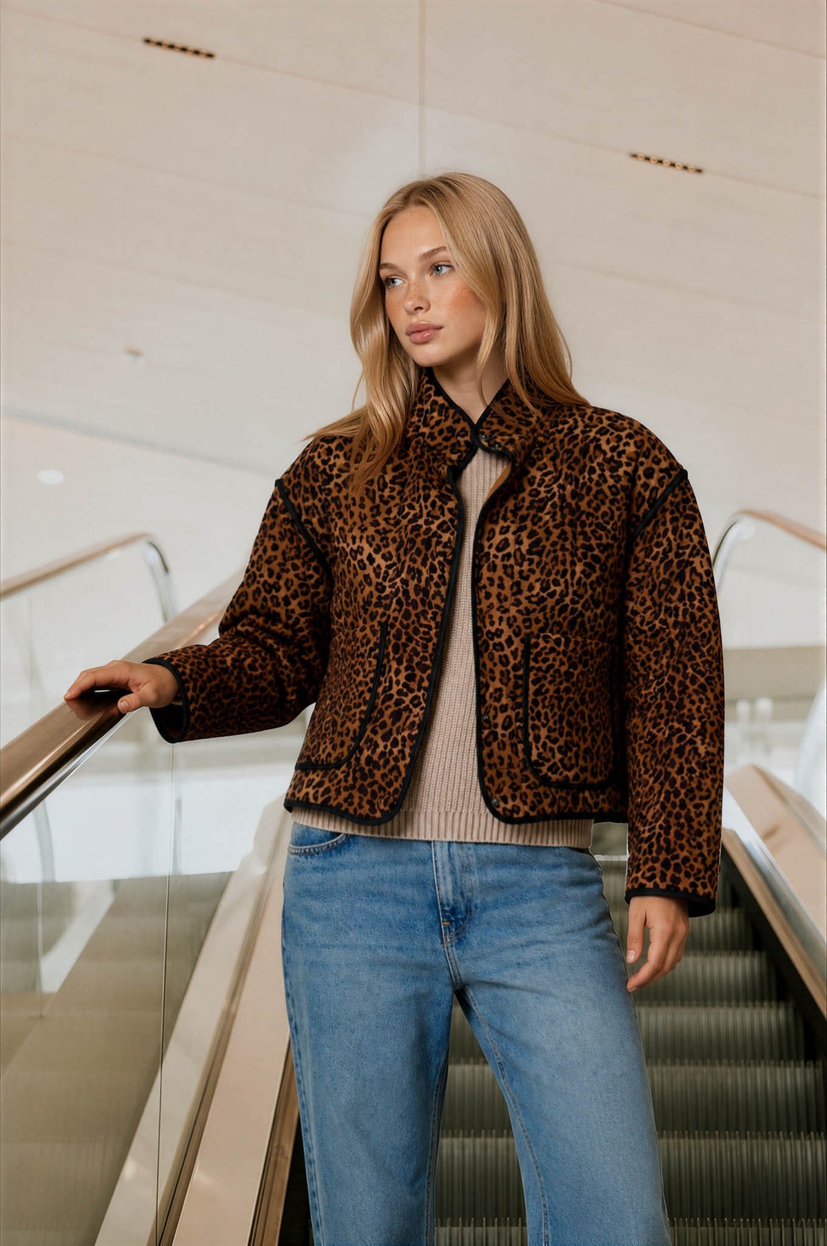 A woman with blonde hair stands on an escalator wearing a brown and black leopard print jacket over a beige ribbed top and blue jeans.