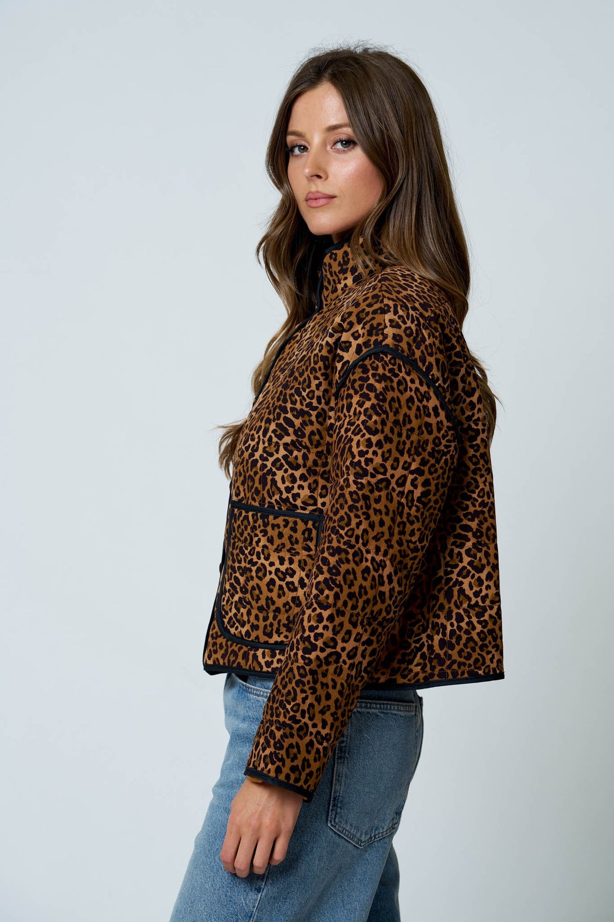 A woman wearing a cropped leopard-print jacket with black trim, paired with blue jeans, photographed against a light gray studio background.