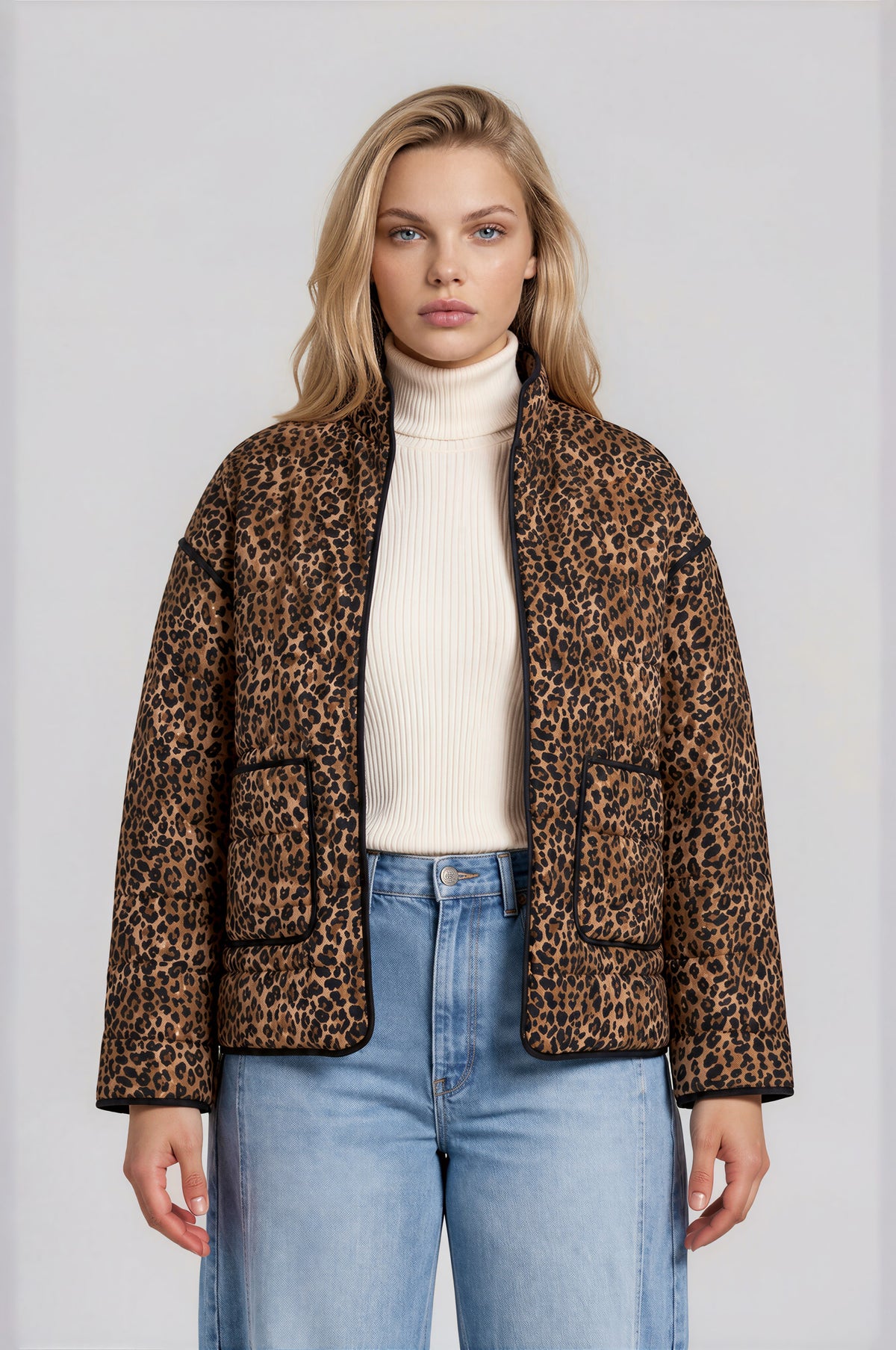 A woman modeling a leopard-print quilted jacket over a cream ribbed turtleneck and light blue jeans, shot against a plain light gray studio background.