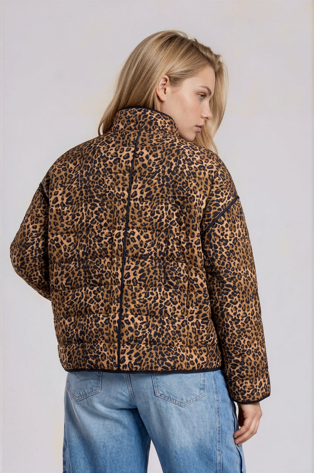 A woman shown from the back wearing a leopard-print puffer jacket and light blue jeans against a plain light gray studio background.