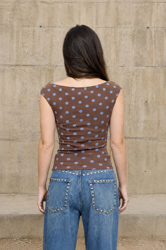 A woman viewed from behind wearing a brown off-the-shoulder top with light blue polka dots and blue jeans, standing against a concrete wall.