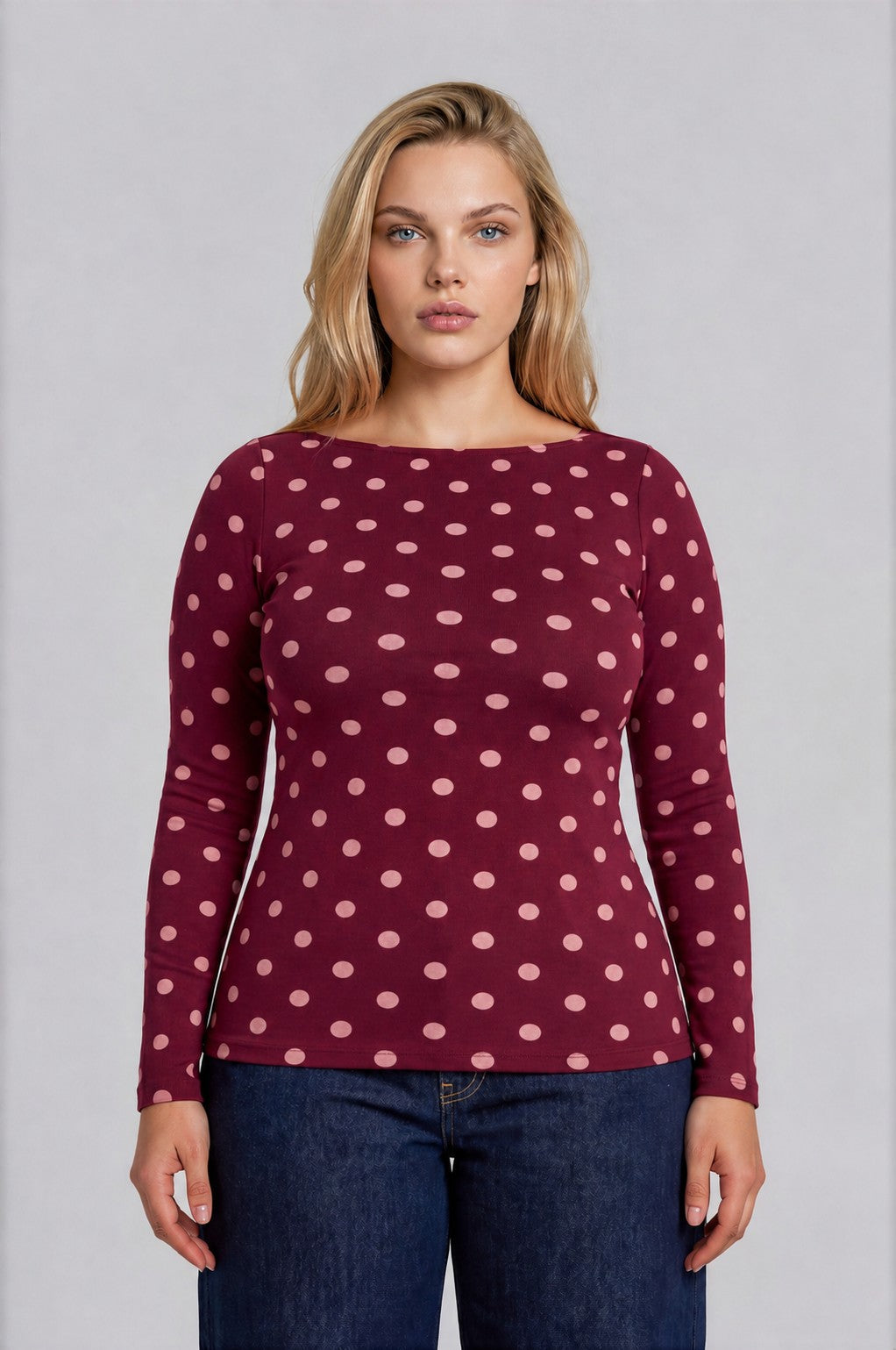 A woman modeling a long-sleeve boat-neck top in burgundy with a light pink polka dot pattern, paired with dark blue jeans, against a solid gray studio background.