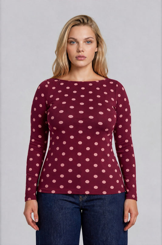 A woman modeling a long-sleeve boat-neck top in burgundy with a light pink polka dot pattern, paired with dark blue jeans, against a solid gray studio background.