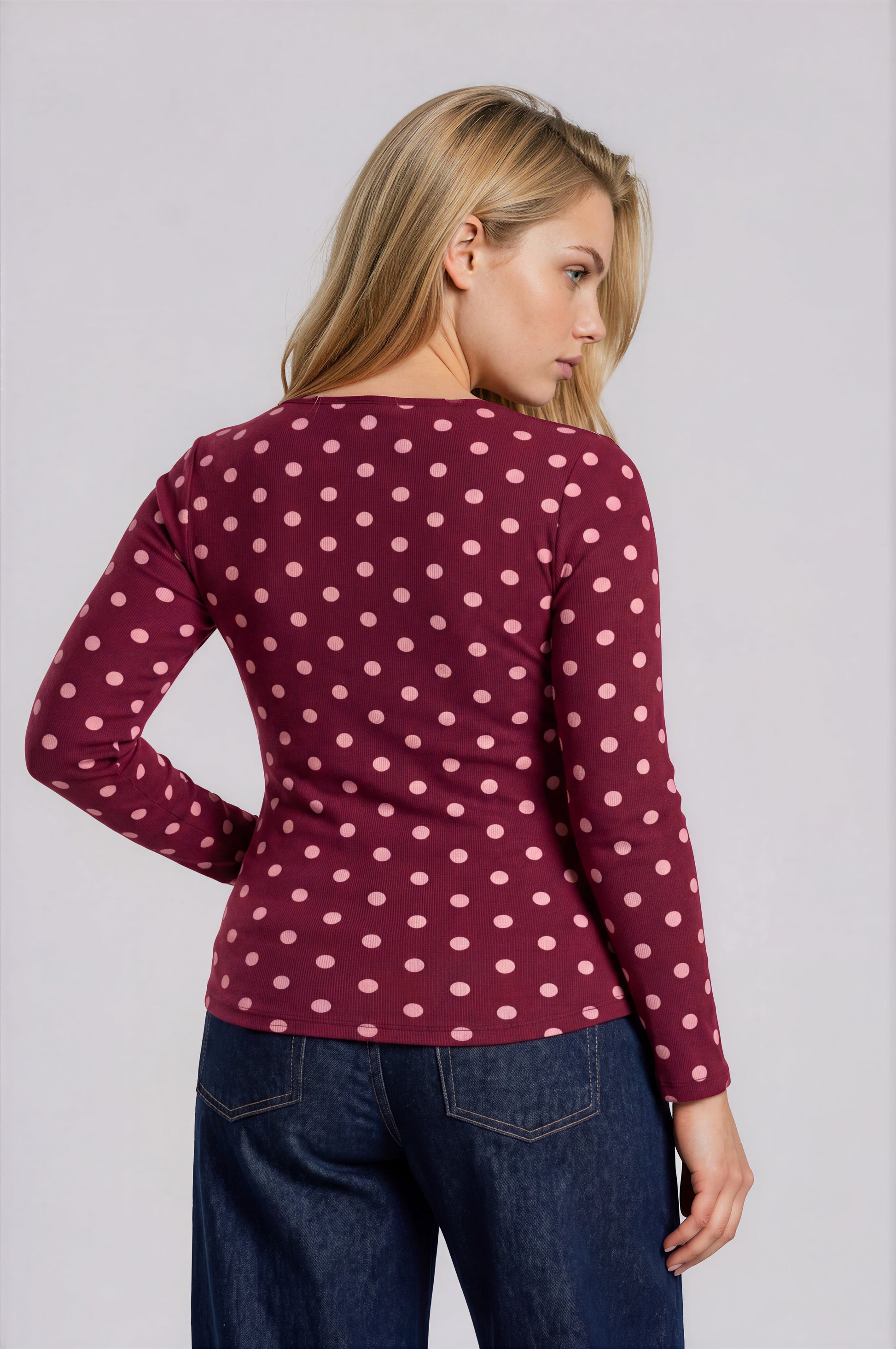 Woman wearing a maroon long-sleeve top with pink polka dots and blue jeans on a light gray background