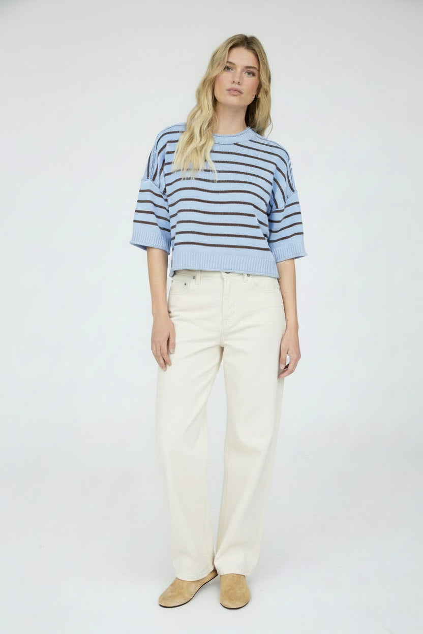 A woman modeling a light blue and brown striped short-sleeve knit top paired with cream denim pants and tan slip-on shoes, against a plain white studio background.