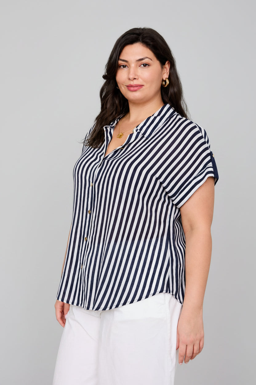 A woman modeling a short-sleeve button-front blouse with vertical navy and white stripes, paired with white pants, against a neutral studio background.