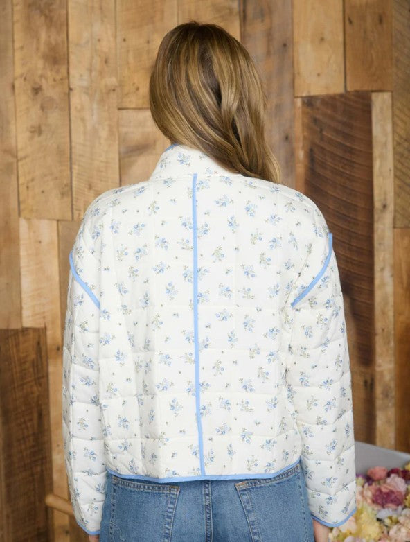 Person wearing a floral quilted jacket with blue piping against a wooden wall.