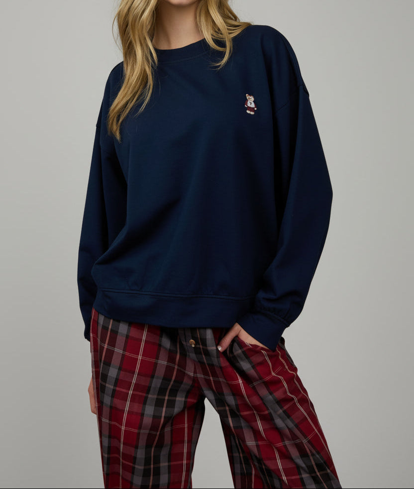 Woman wearing a navy sweatshirt with a logo and plaid pants against a wooden paneled wall.