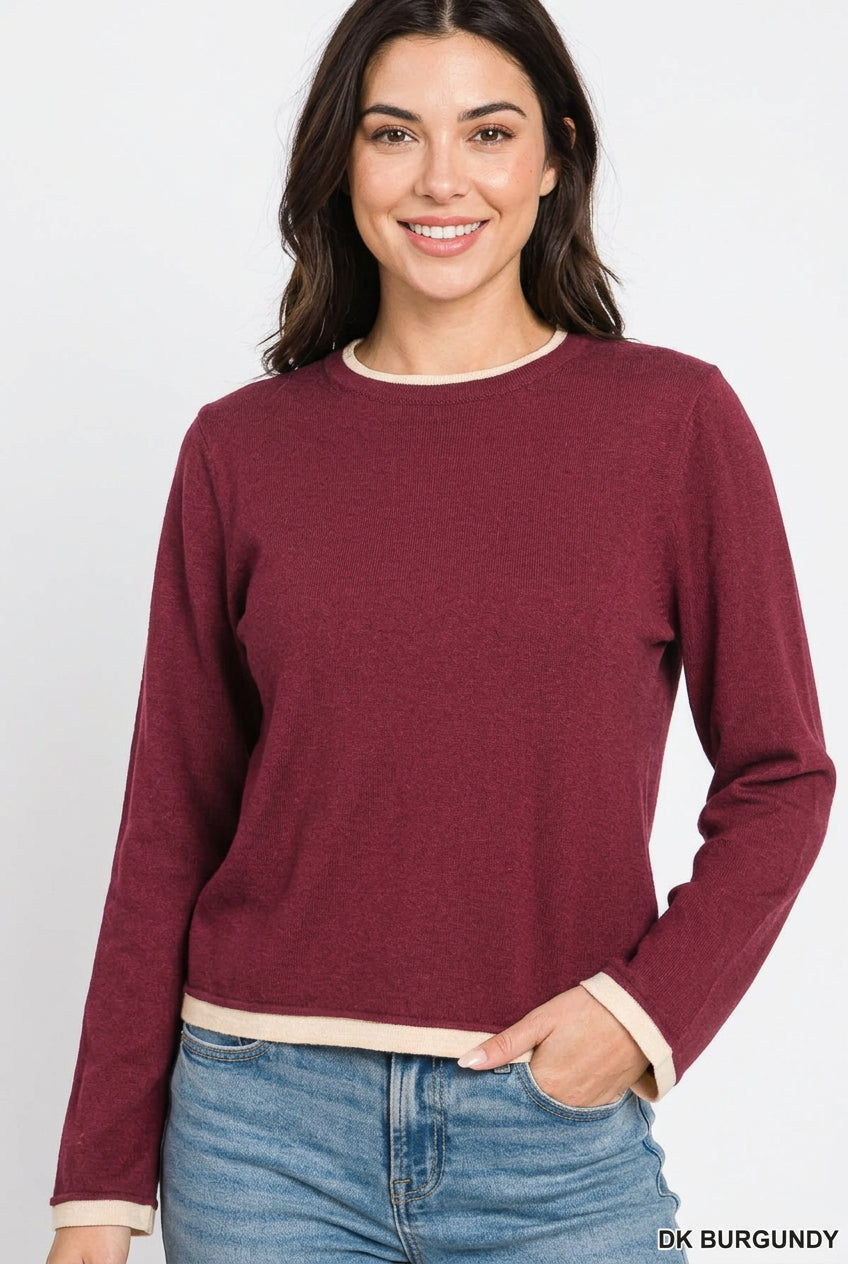 A woman wearing a long-sleeve crewneck sweater in burgundy with cream trim at the collar, hem, and cuffs, paired with blue jeans against a white background.
