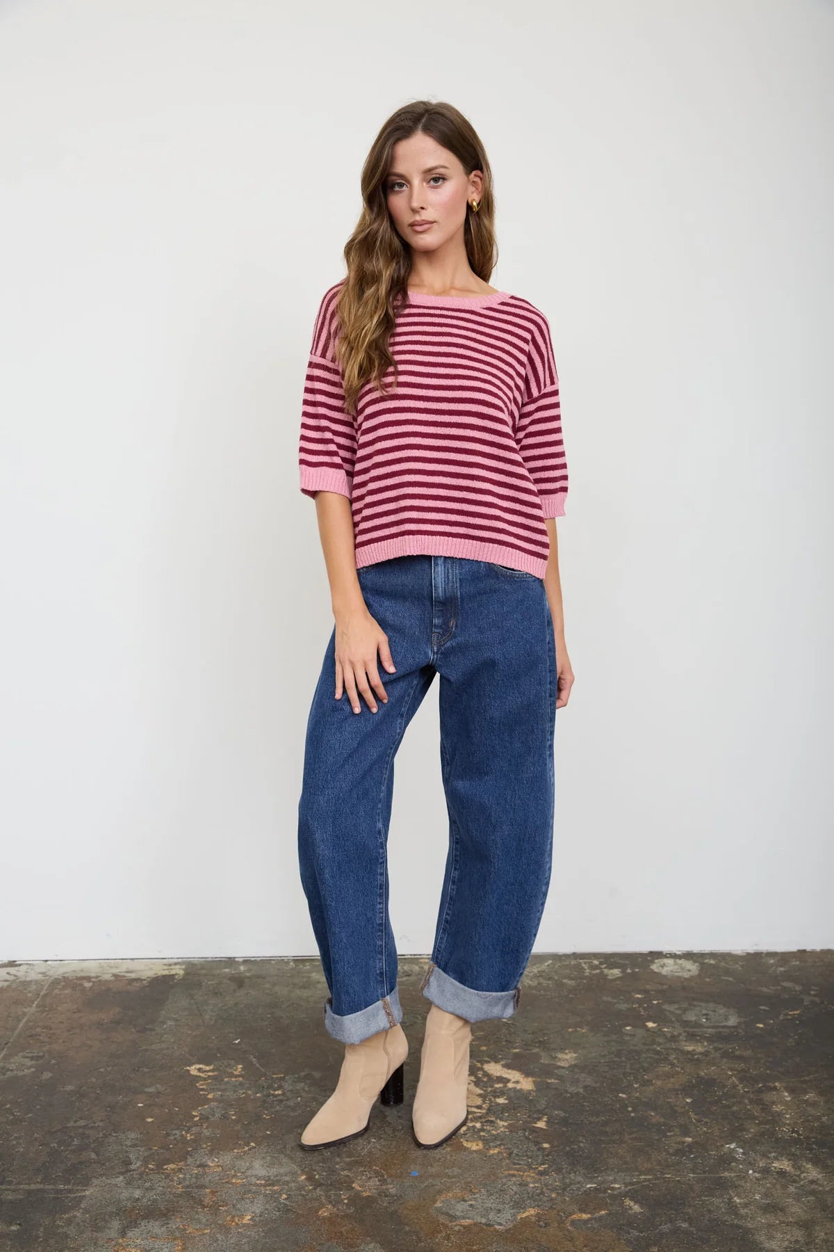A woman modeling a pink and burgundy striped knit top with three-quarter sleeves, paired with cuffed blue jeans and beige ankle boots, standing against a white wall.