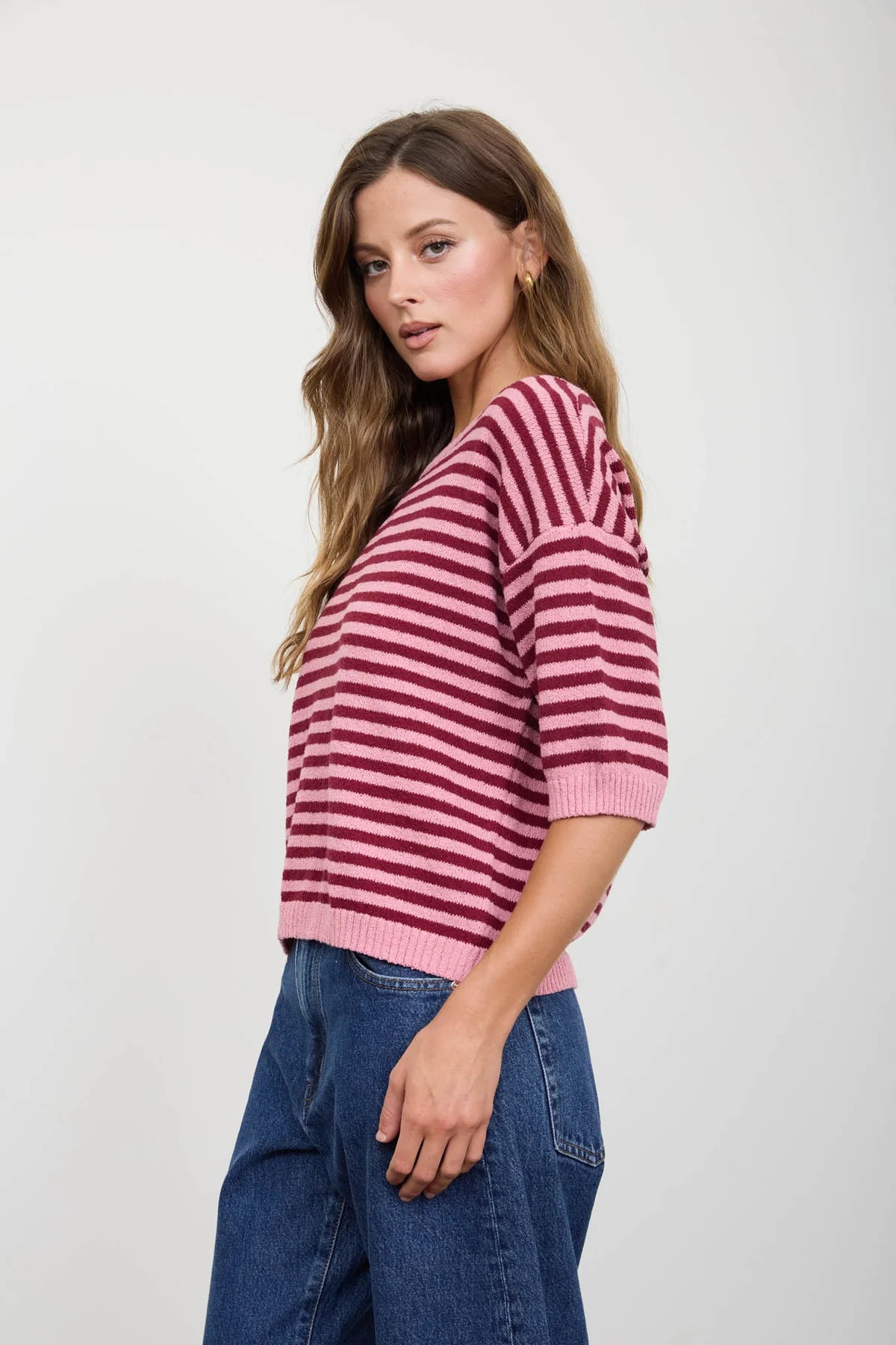 A woman wearing a pink and burgundy striped knit sweater with three-quarter sleeves and blue jeans, photographed against a plain light gray studio background.