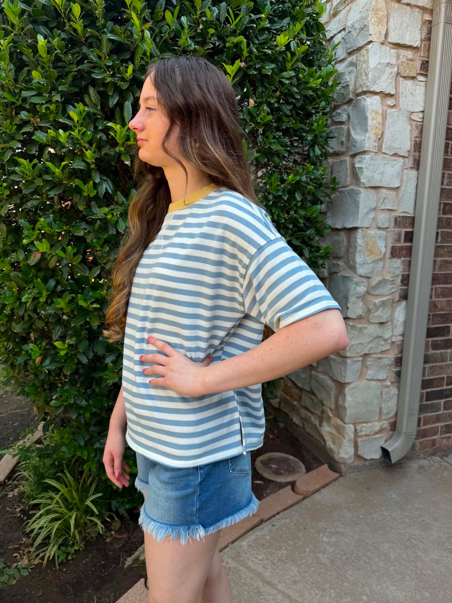A woman wearing a light blue and white striped short-sleeve t-shirt and denim shorts, standing outdoors next to a stone wall and green hedge.