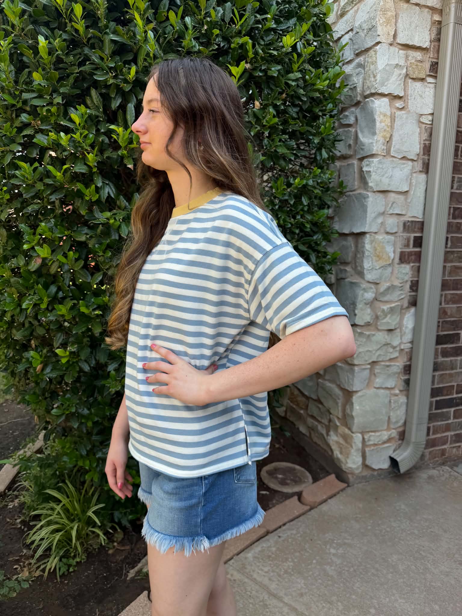 A woman wearing a light blue and white striped short-sleeve t-shirt and denim shorts, standing outdoors next to a stone wall and green hedge.