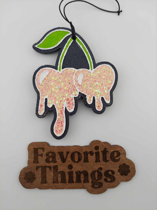 A handmade freshie in the shape of a pink glittering cherry with white specks, hanging on a black elastic string with a label reading 'Favorite Things'.