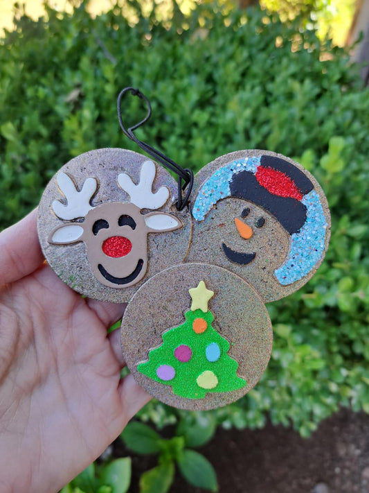 Three Christmas-themed cookie freshies shaped like a reindeer, snowman, and tree held by a hand with a green outdoor background.