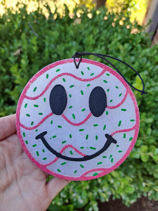 Decorative circular freshie with a smiley face design held in front of green foliage.