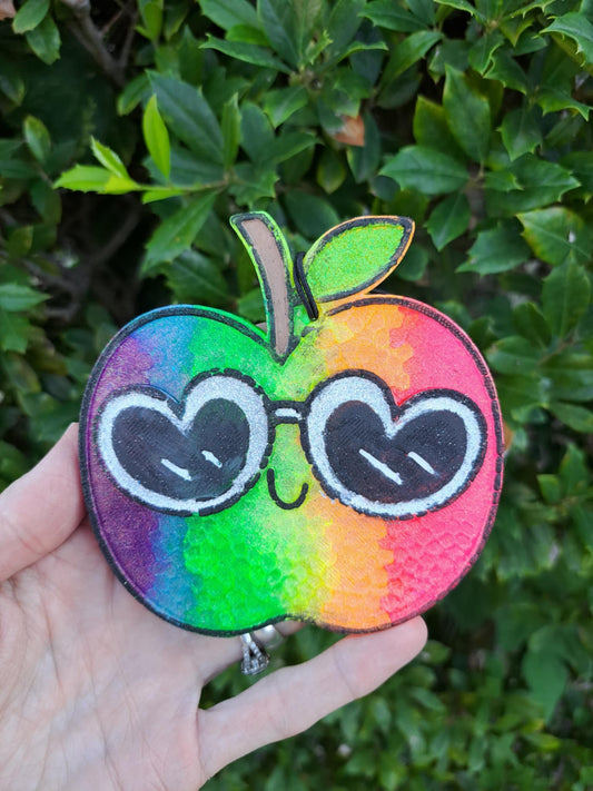 Colorful apple freshie with heart sunglasses held in front of green foliage.