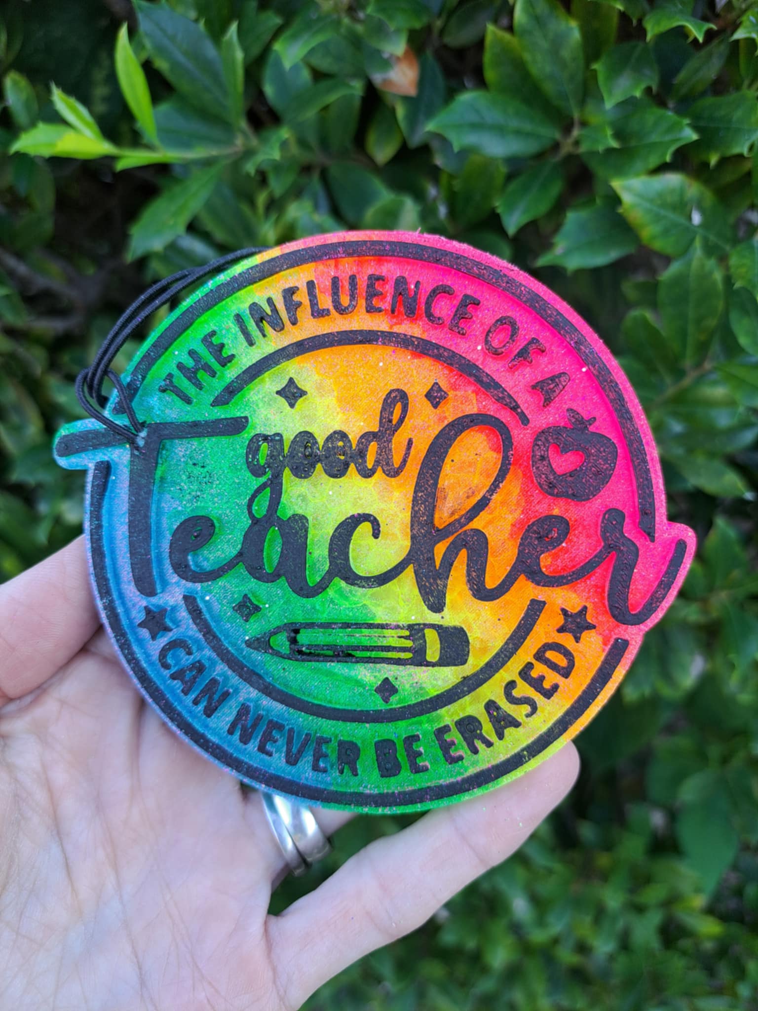 Colorful freshie with black text 'good teacher' held by a hand against a green leafy background