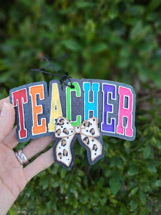 Colorful 'TEACHER' freshie with a leopard bow held in front of a green leafy background