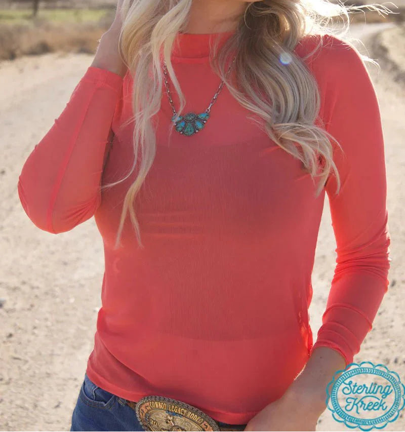 Woman wearing a red long-sleeve top with a necklace, standing in a desert-like setting.