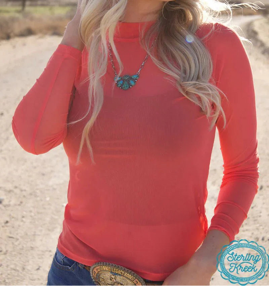 Woman wearing a red long-sleeve top with a necklace, standing in a desert-like setting.