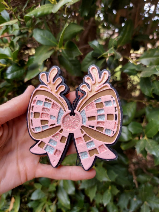 A pink and beige disco bow air freshener with antlers, held in a hand against a backdrop of green foliage.