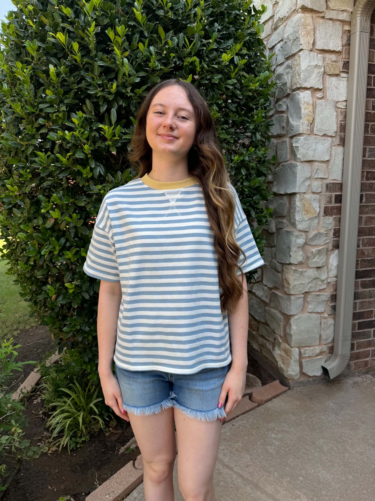A young woman stands outdoors in front of a stone wall and green hedge, wearing a light blue and white striped short-sleeve t-shirt with a yellow collar trim and denim shorts.
