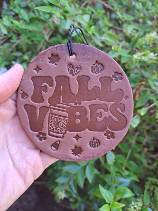 Round brown ornament with 'Fall Vibes' text and fall-themed designs held against a green leafy background.