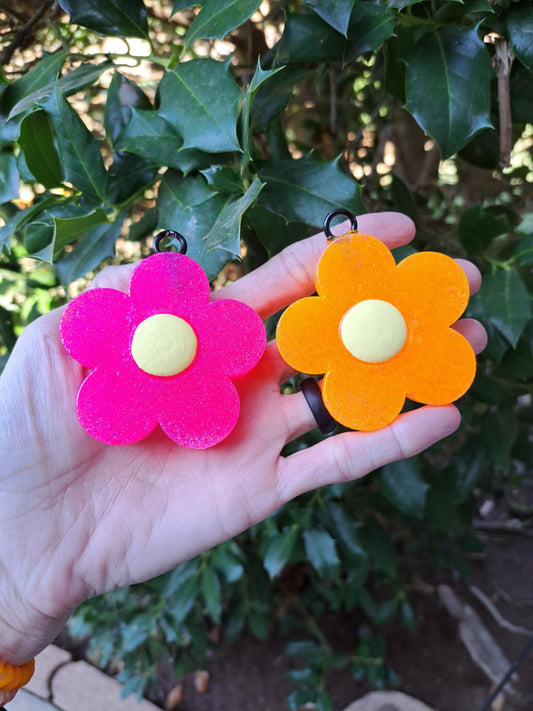Two flower-shaped vent clip fresheners held in hand, one pink and one orange, with green leaves in the background.