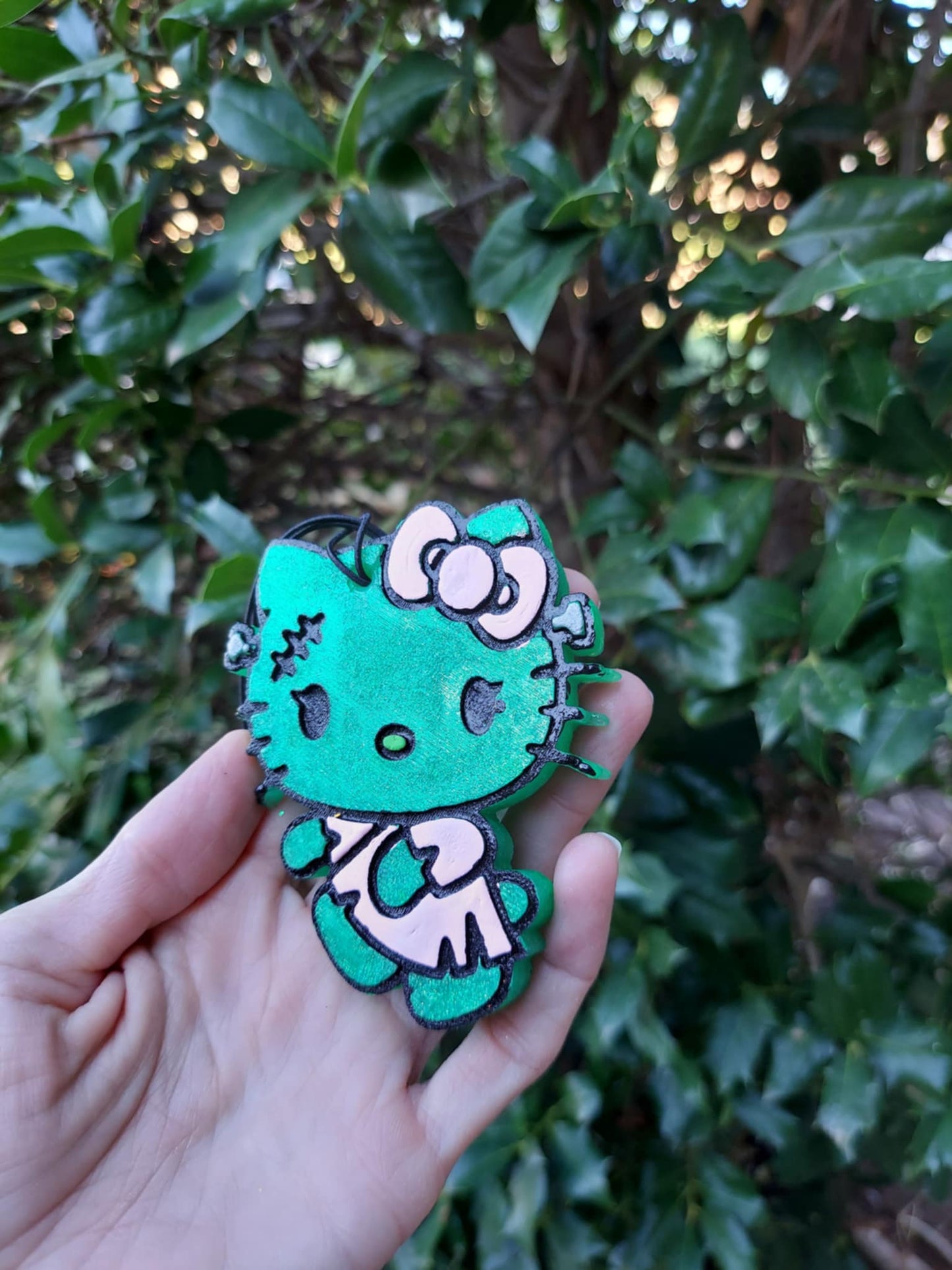 Hand holding a green air freshener shaped like a stylized kitty with a floral design and black elastic string.