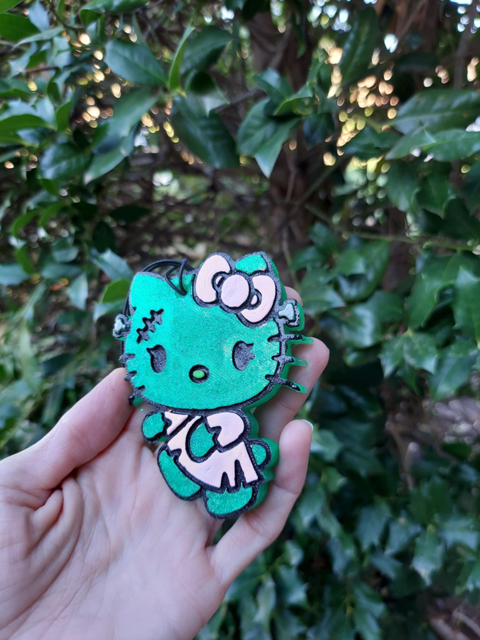 Hand holding a green air freshener shaped like a stylized kitty with a floral design and black elastic string.