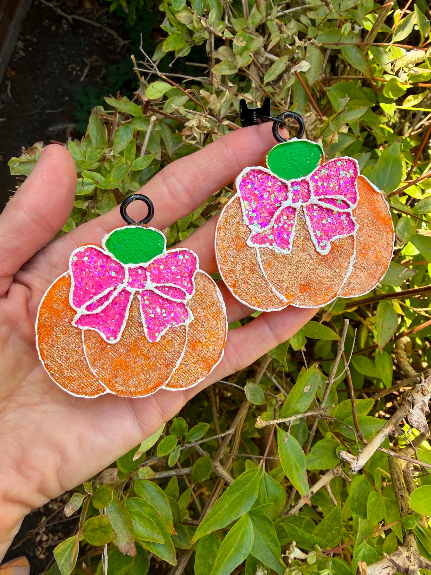 Two glittery orange pumpkin-shaped vent clip freshies with pink bows and green tops, held in a hand against a background of green outdoor foliage.