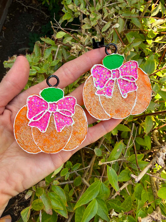 Two glittery orange pumpkin-shaped vent clip freshies with pink bows and green tops, held in a hand against a background of green outdoor foliage.