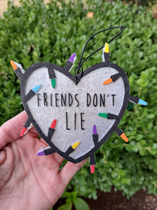 Heart-shaped ornament with 'Friends Don't Lie' text and colorful Christmas Lights, held against a green bush background.