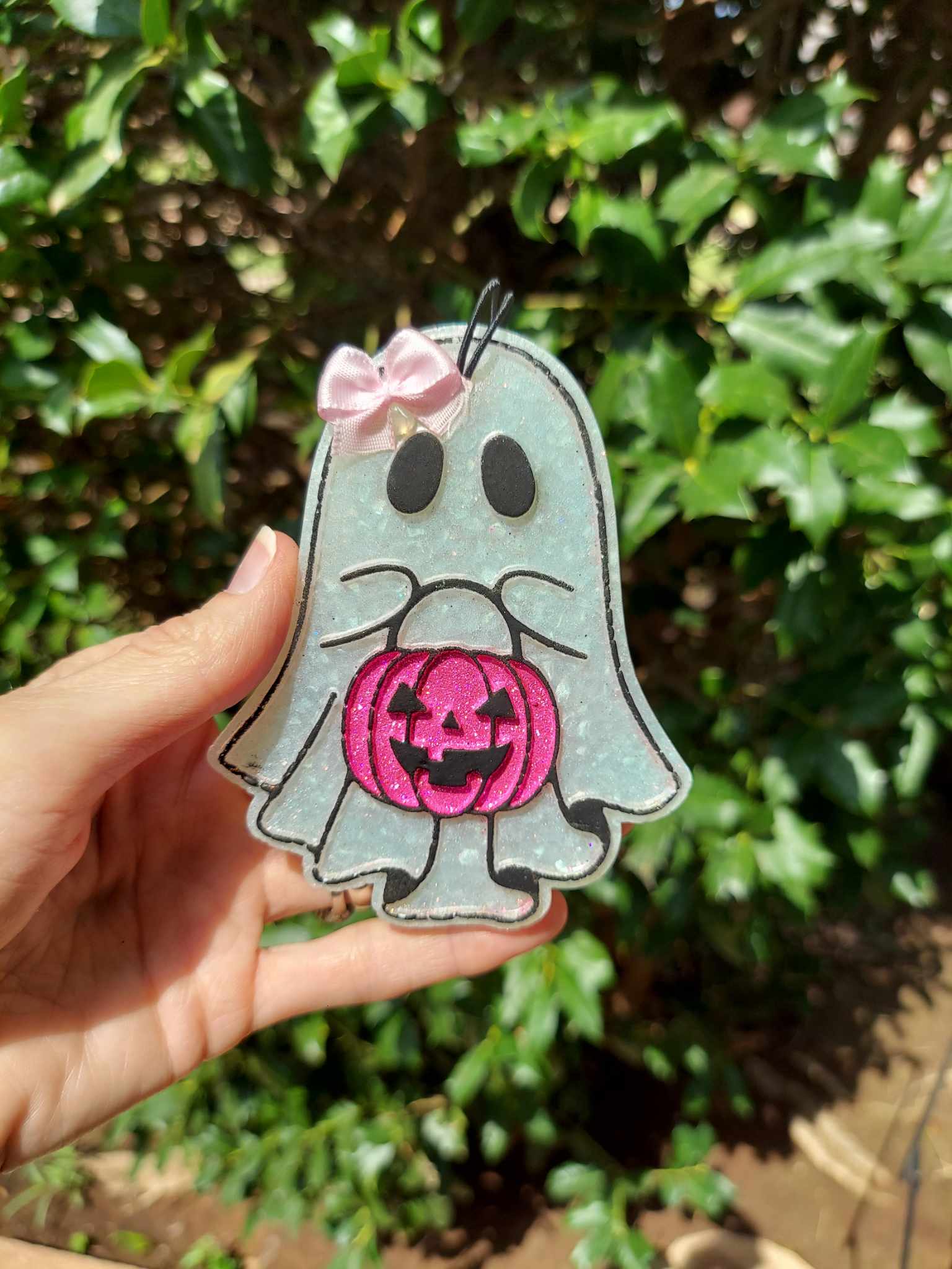 A hand holding a small air freshener shaped like a ghost with a pink pumpkin, against a backdrop of greenery.