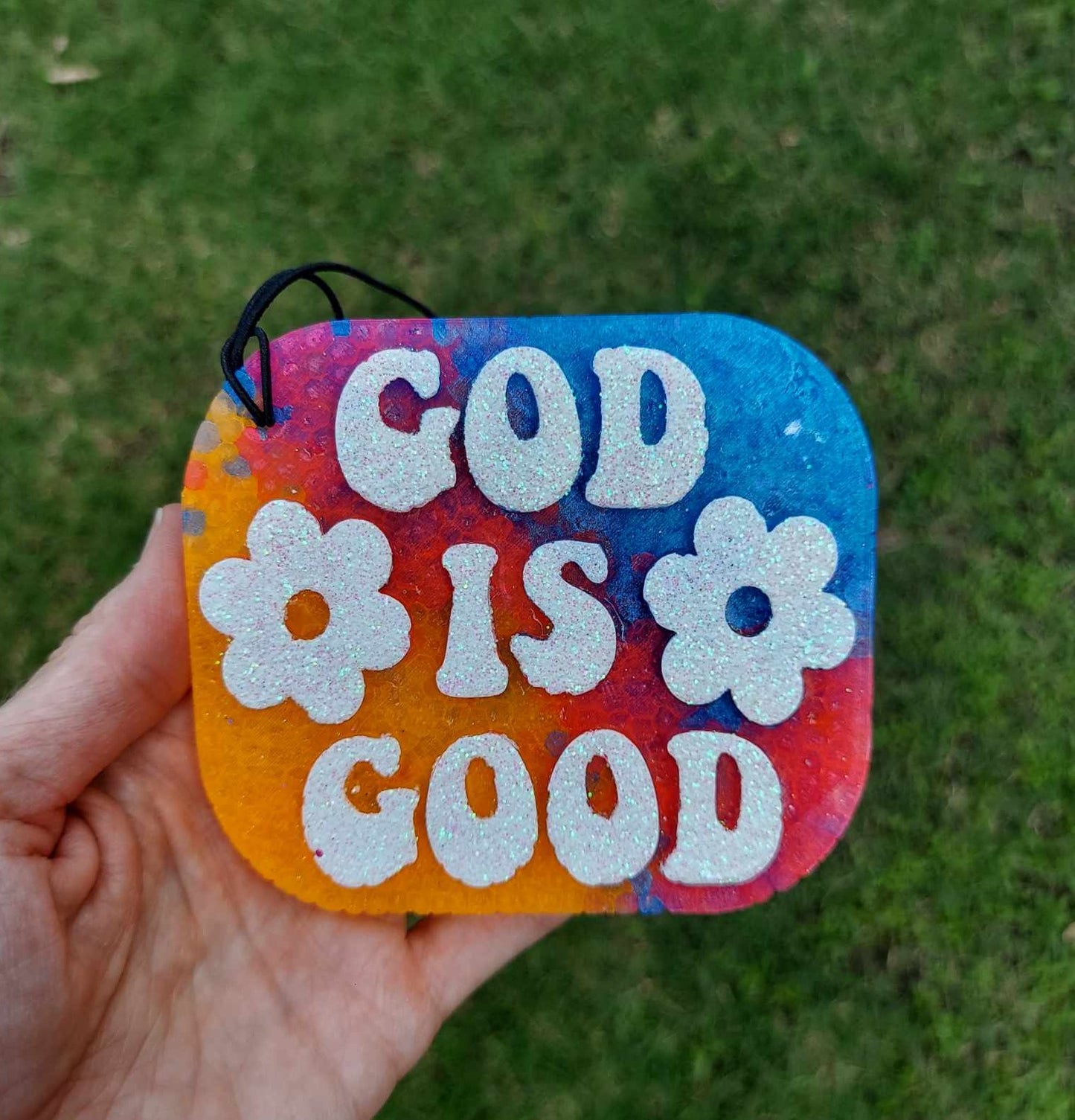 A hand holding a tie-dye air freshener with the text 'GOD IS GOOD' in white on a colorful background, with a black elastic string.