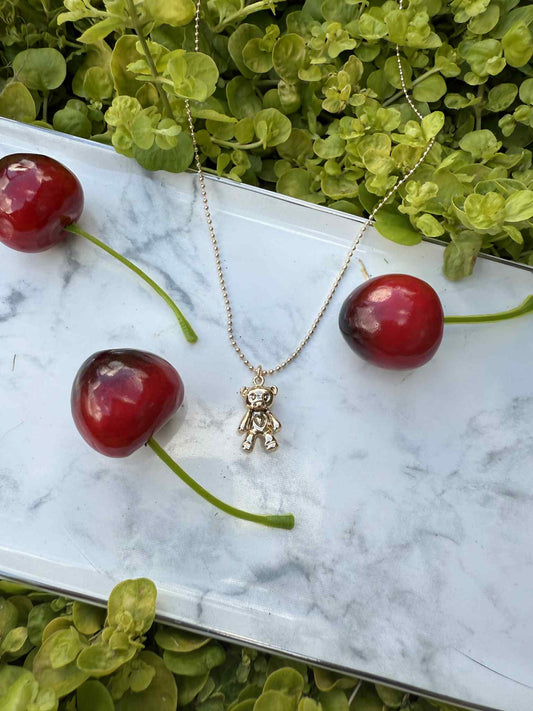 Gold teddy bear necklace on a marble surface with cherries and green leaves.
