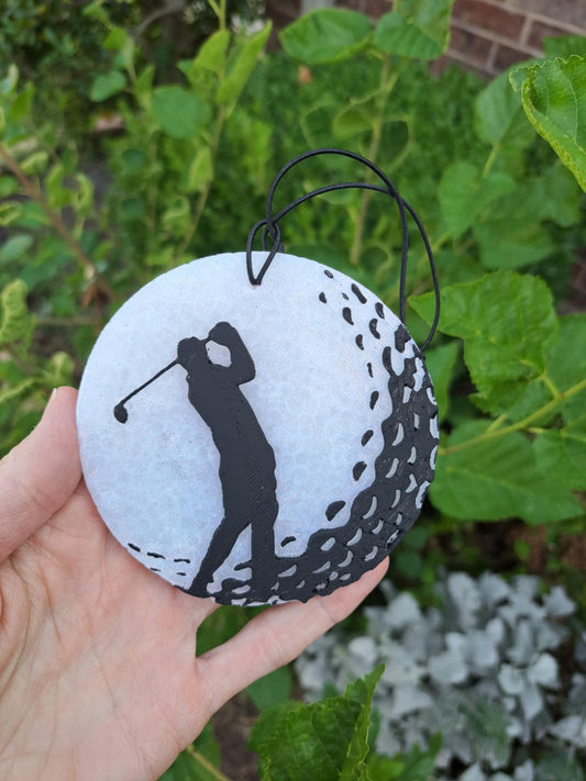 Golfer silhouette on golf ball shaped air freshener with a white background held in front of green foliage.