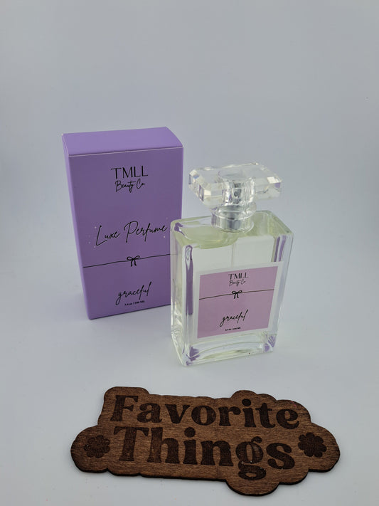 A glass bottle of 'Luxe Perfume - Graceful' with a purple label, next to its purple box packaging, on a wooden surface with a 'Favorite Things' plaque.