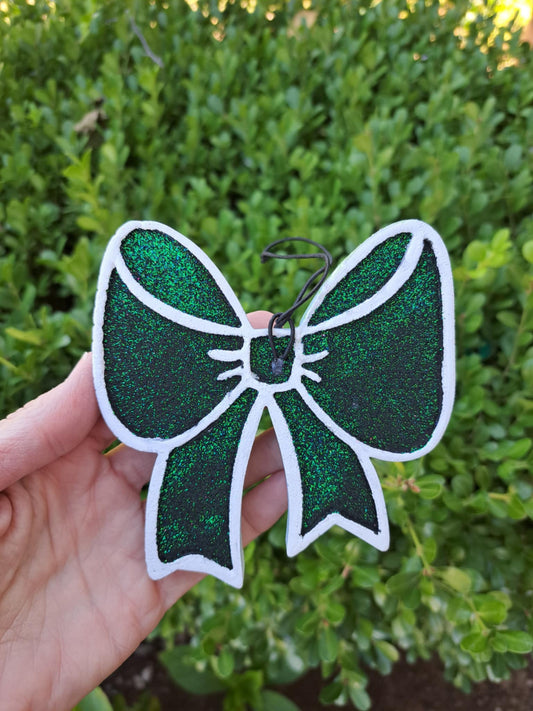 Green glittery bow freshie held by a hand with a blurred green leafy background