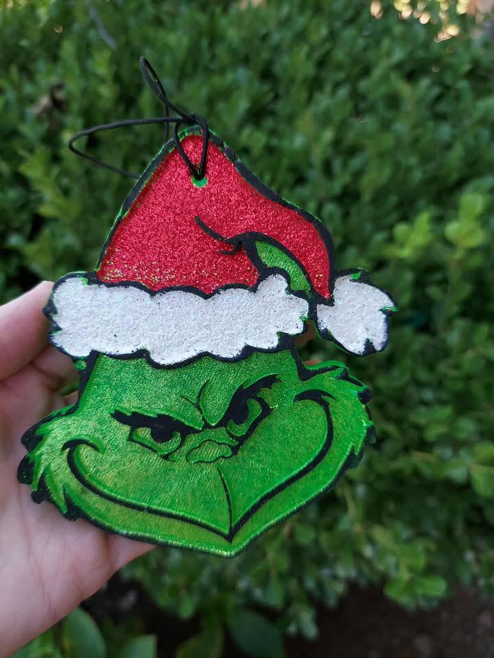 Decorative freshie shaped like the Grinch with a red Santa hat held against a green leafy background.