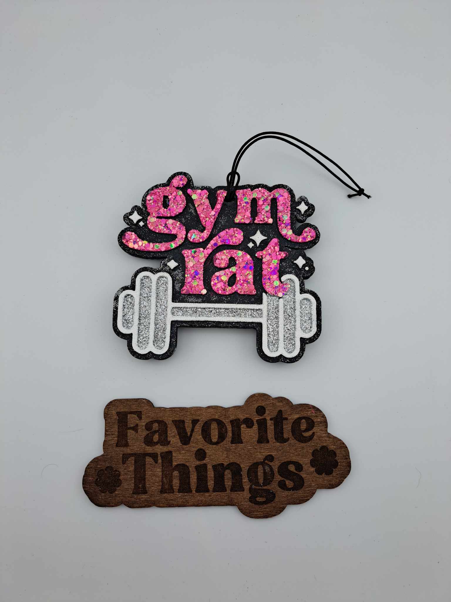 A handmade, silver and pink air freshener with the text 'Gym Rat' and a barbell symbol on top, and a brown tag reading 'Favorite Things' at the bottom.