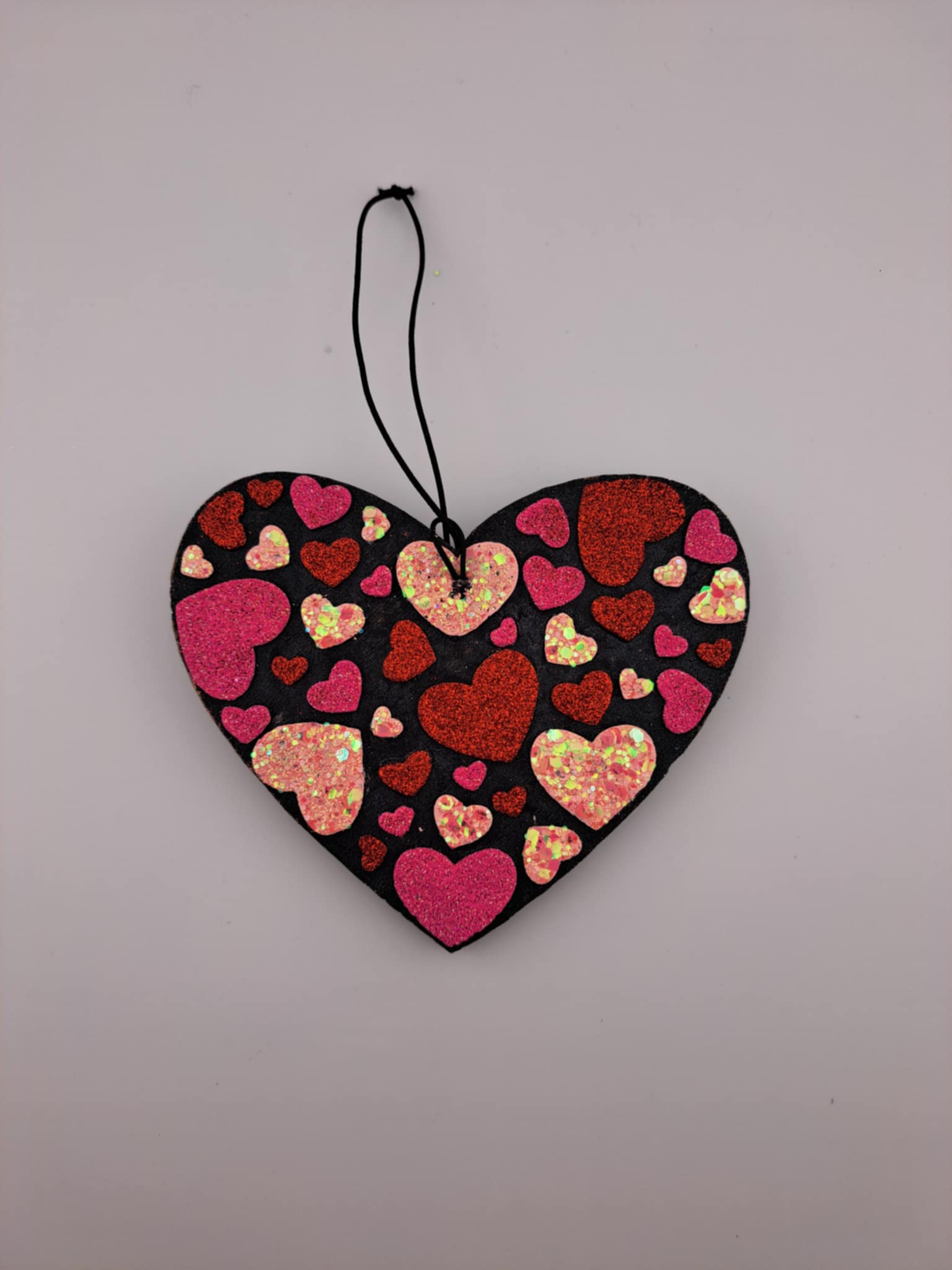 A heart-shaped air freshener with a pattern of multicolored hearts on a black background, hanging on a black elastic string.