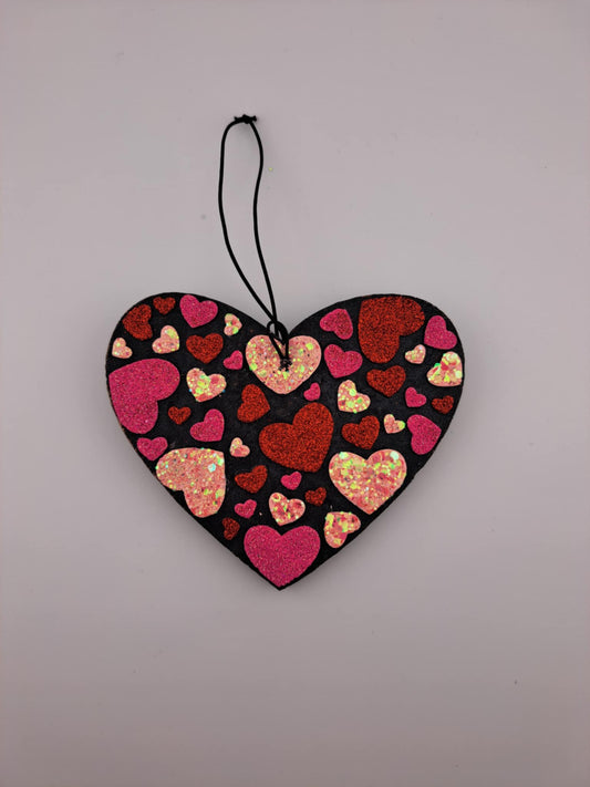 A heart-shaped air freshener with a pattern of multicolored hearts on a black background, hanging on a black elastic string.