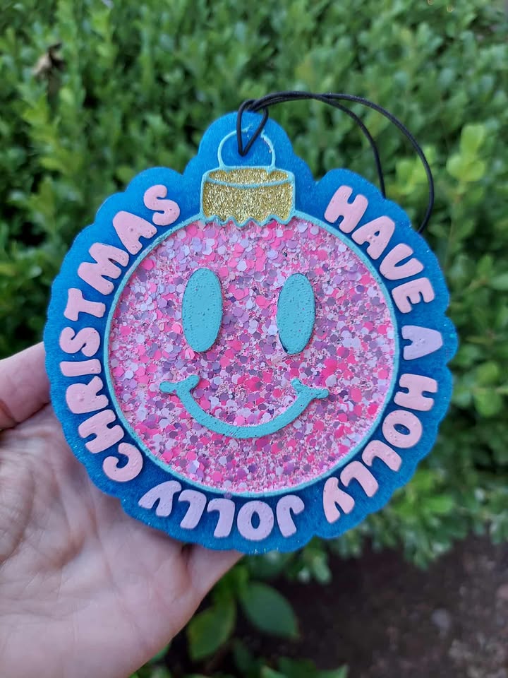 Decorative freshie with pink glittery Christmas bulb and text 'Have a Holly Jolly Christmas' held against a green bush background.