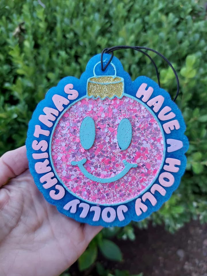 Decorative Christmas ornament freshie with pink glittery ball and blue text held by a hand against a green bush background.